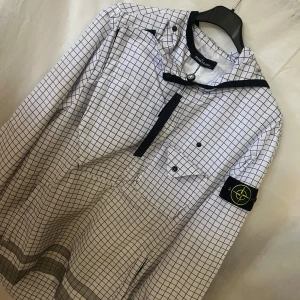 Vit rutig vindjacka från Stone Island - Vit vindjacka från Stone Island med svart rutmönster, huva och dragkedja framtill. Jackan har klassisk logotyp-patch på ärmen, bröstficka med knapp och detaljer i svart. Materialet är polyester och jackan har en loose passform.