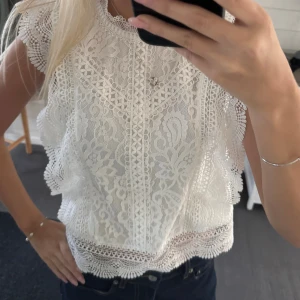 Vit spetsblus med volangdetaljer☁️ - Superfin vit blus i spets med vackra broderade mönster och volangdetaljer längs ärmhål och nederkant. Blusen är ärmlös och har en hög halsringning med dekorativa kanter. Perfekt för dig som gillar romantisk och feminin stil.