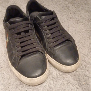 Svarta sneakers med broderad fågel - Svarta sneakers från Axel Arigato i skinn med broderad färgglad fågel på sidan. Skorna har svarta snören, vit sula och klassisk rund tå. Perfekta för dig som vill ha en stilren men ändå unik look.