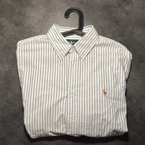 Randig skjorta från Ralph Lauren - Klassisk skjorta från Ralph Lauren i vitt och ljusblått med smala vertikala ränder. Custom fit-modell med lång ärm, knappar framtill och broderad logga på bröstet. Tillverkad i mjuk bomull, perfekt för en clean och stilren look. Storlek L men passar mer som M enligt mig. 