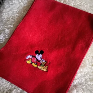 Röd fleecehalsduk med Disney-brodyr - Helt ny, röd halsduk i mjuk fleece med broderat motiv av Musse Pigg och Pluto. Perfekt accessoar för dig som gillar klassiska Disney-karaktärer och vill ha något färgstarkt och mysigt runt halsen. Halsduken är rektangulär och har en enkel, stilren design. Mjuk och skön material. 