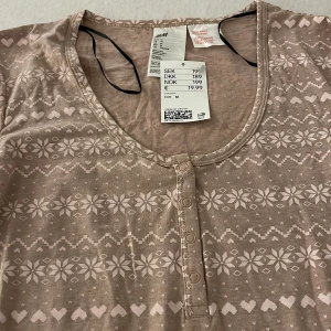 Beige långärmad tröja från H&M - Supersöt beige långärmad tröja från H&M med ljusa vintermönster av snöflingor och hjärtan. Rund halsringning och knappdetaljer framtill. Perfekt för mysiga dagar och vintersäsongen.