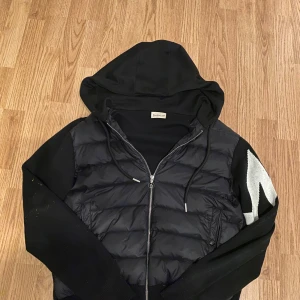 Moncler cardigan  - Säljer min svarta Moncler huvjacka som bara är använd ett fåtal gånger och fortfarande i perfekt skick. Den har en quiltad front med vaddering som håller värmen bra, medan ärmar och baksida är i mjukt stickat material för en avslappnad men exklusiv look. Stor och tydlig Moncler-logga på ärmen som ger ett stilrent statement.  Alla originaldetaljer finns kvar – inklusive Moncler-graverade metalltips på snörena. NFC-taggen och QR-koden fungerar precis som de ska.  Pris kan diskuteras vid seriöst in
