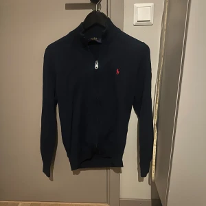 Mörkblå kofta från Polo Ralph Lauren - Mörkblå kofta från Polo Ralph Lauren med dragkedja framtill och klassisk röd broderad logga på bröstet. Tröjan är ribbstickad vid kragen och ärmarna, och är tillverkad i mjuk bomull. Perfekt för lager-på-lager och snygg till jeans.