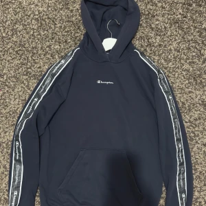 Mörkblå Champion hoodie med logoband - En marinblå snygg Champion hoodie där det står champion på sidan av armarna. Har använts men väldigt sparsamt.   Några funderingar eller vill ha fler bilder, hojta bara till!