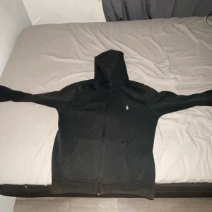 Svart hoodie från Polo Ralph Lauren - Svart hoodie från Polo Ralph Lauren med klassisk vit broderad logga på bröstet. Tröjan har dragkedja framtill, huva och känguruficka. Tillverkad i mjuk bomullsmix, perfekt för chill dagar. Enkel och stilren design som funkar året runt.