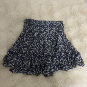 Blommig volangkjol - Supersöt kjol med vitt blommönster och volangdetaljer. Högmidjade men går att vikas ner. Smock i midjan för extra komfort och en flowy känsla. 