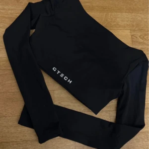 Svart långärmad träningstopp GTECH - Säljer en svart långärmad croppad träningstopp från GTECH. Toppen har en tight passform och är tillverkad i ett stretchigt syntetmaterial som passar perfekt för gym eller löpning. Snygg och stilren med logga framtill.