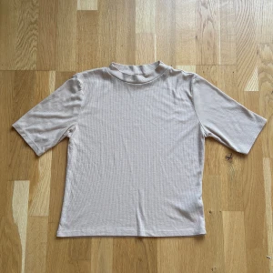 Beige ribbad polotopp från H&M - Säljer en beige ribbad polotopp från H&M Divided. Toppen har korta ärmar och en högre krage som ger en clean look. Materialet är mjukt och stretchigt med 64% polyester, 32% viskos och 4% elastan. Perfekt att styla med jeans eller kjol.