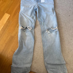 highwaist ljusblå destroyed jeans - Säljer ett par 90-talsinspirerade ljusblå jeans med hög midja och slitna hål över knäna. Jeansen har en rak passform och klassisk femficksdesign. Perfekta för dig som gillar en avslappnad och trendig look.