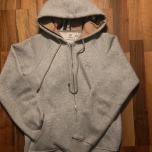 Grå hoodie från Burberry med dragkedja - Säljer en grå hoodie från Burberry med klassiskt rutigt foder i huvan och broderad logga på bröstet. Tröjan har dragkedja framtill, snörning i huvan och två fickor. Perfekt för dig som gillar stilrena och exklusiva plagg.