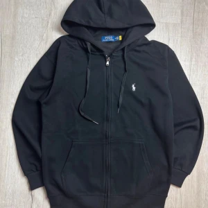 Svart hoodie från Polo Ralph Lauren - Svart hoodie från Polo Ralph Lauren med klassisk broderad logga på bröstet. Tröjan har huva med snörning, dragkedja framtill och en stor magficka. Tillverkad i 100% bomull för skön och avslappnad känsla. Perfekt för chill dagar och streetwear.