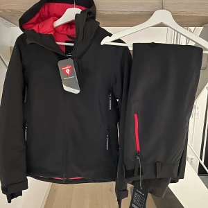 Svart skiddress från J.Lindeberg - Svart skiddress från J.Lindeberg med cerise insida och detaljer, huva och flera dragkedjor.  Primaloft-teknologi och har en clean, sportig look. Perfekt för vintern och skidbacken, snygg passform. Nytt med prislapparna kvar. Jacka stl S byxa stl M