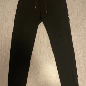 Svarta träningsbyxor från BOSS - Svarta träningsbyxor från BOSS med broderad logga på bakfickan och snygga metallringar vid fickorna. Byxorna har snörning i midjan och en rak, normal passform. 