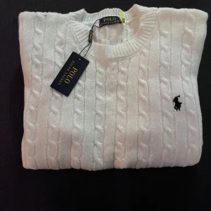 Vit kabelstickad tröja Polo Ralph Lauren - Snygg vit kabelstickad tröja från Polo Ralph Lauren med klassisk svart logga på bröstet. Rund hals och långa ärmar, ribbade muddar och mjukt material som känns lyxigt. Perfekt för dig som gillar stilrena och tidlösa plagg.