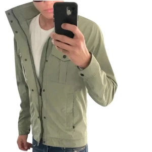 Grön vindjacka från J.Lindeberg - Stilren ljusgrön vindjacka från J.Lindeberg med hög krage, dragkedja och tryckknappar framtill. Jackan har flera fickor och diskret logga på ärmen. Perfekt för vår och höst när du vill ha en lätt och snygg jacka.