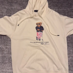 Vit hoodie Polo Bear Ralph Lauren - Säljer en vit hoodie från Polo Ralph Lauren med ikoniska Polo Bear-trycket på bröstet. Tröjan har huva med snörning, känguruficka och en avslappnad passform. Perfekt för dig som gillar streetstyle och klassiska märken. KNAPPT ANVÄND!!!