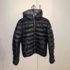 Svart dunjacka från Colmar med huva - Säljer en svart dunjacka från Colmar med huva och två fickor med dragkedja. Jackan har ett stilrent, quiltat mönster och är perfekt för kalla dagar. Den har hel dragkedja framtill och en snygg logga på ärmen. Passformen är normal och jackan är riktigt lätt och skön. Storlek 42 vilket motsvarar xs-s