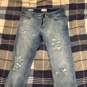 Jack & Jones ljusblå skinny jeans - Säljer ett par ljusblå skinny fit jeans från Jack & Jones, modell Liam. Jeansen har slitna detaljer och hål på benen för en cool, trendig look. Klassisk femficksdesign och knappgylf. Perfekta för dig som gillar smal passform och streetstil.