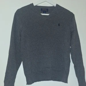 Grå v-ringad tröja Polo Ralph Lauren - Grå v-ringad tröja från Polo Ralph Lauren med mörkblå broderad logga på bröstet och svarta mocka-lappar på armbågarna. Tröjan är stickad och har långa ärmar med ribbade muddar. Perfekt för dig som gillar klassisk stil med en twist.