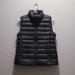 Svart dunväst från Uniqlo - Säljer en svart, quiltad dunväst från Uniqlo Ultra Light Down. Västen är lätt, har hög krage och dragkedja framtill. Perfekt för lager-på-lager och enkel att bära med sig. Klassisk och stilren design utan ärmar.