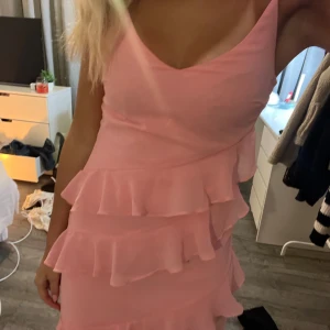 Lång rosa balklänning med volanger - Säljer en lång, ljusrosa klänning med smala axelband och v-ringad hals. Klänningen har flera lager med volanger som ger en snygg och lekfull look. Materialet känns lätt och flowy, perfekt för student eller bal. Passformen är normal och klänningen är ärmlös. Någon fläck på benet men syns knappt 