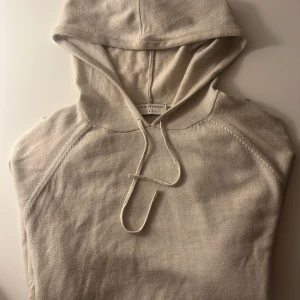 Beige hoodie från Tiger of Sweden - Snygg beige hoodie från Tiger of Sweden med klassisk huva och dragsko. Tröjan är i mjuk merinoull och har långa ärmar med raglanskärning. Perfekt för en chill och stilren look.