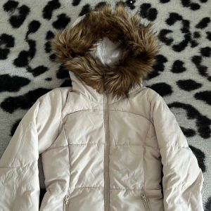 Beige pufferjacka med fuskpäls från H&M - Säljer en beige pufferjacka från H&M Divided med huva och fuskpäls. Jackan har dragkedja framtill och två sidofickor med dragkedja. Perfekt för kalla dagar och har en snygg, enkel design som funkar till många stilar.