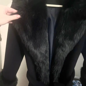 Lyxig mörkblå Ivyrevel-kappa med fuskpäls – perfekt till vintern & hösten - Säljer min fantastiskt fina mörkblå kappa från Ivyrevel – en riktig drömkappa som både känns exklusiv och elegant. Den har mjuk fuskpäls både på kragen och ärmarna, vilket ger den en lyxig och trendig look. Pälsen går dessutom ner längs kappans framsida, vilket gör den extra iögonfallande och varm.  Kappan är lång, mysig och figursydd med ett band i midjan som framhäver siluetten på ett smickrande sätt. Perfekt att bära under hösten och vintern, och varför inte till nyår för en riktigt glamorös 