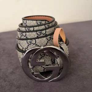 Gucci GG canvasbälte med silverdetalj - Snyggt bälte från Gucci i beige canvas med klassiskt GG-mönster i mörkblått. Stor rund spänne i silverfärgad metall med dubbla G-loggan. Insidan är i ljust skinn. Perfekt accessoar för att lyfta din outfit med en lyxig touch.