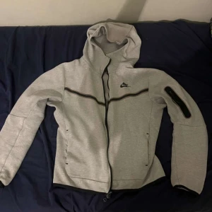 Grå Nike vindjacka med huva - Säljer en grå vindjacka från Nike med huva och hel dragkedja framtill. Jackan har svarta detaljer längs bröstet och på ena ärmen, samt praktiska fickor. Perfekt för dig som gillar sportig och clean stil.