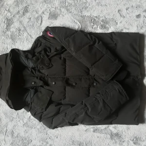 Svart dunjacka från Canada Goose M - Svart dunjacka från Canada Goose i storlek M. Jackan har huva, quiltad design och snygga detaljer med knappar framtill. Märkets klassiska patch syns på axeln. Fylld med dun och fjädrar för extra värme, perfekt för kalla dagar. Det ända felet med jackan är märket på dragkedjan. Annars är den perfekt till vinter tiderna!