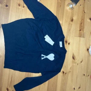 Marinblå stickad tröja från Ami - Snygg marinblå stickad tröja från Ami Alexandre Mattiussi med vit hjärtlogga på bröstet. Tröjan har ribbade muddar vid hals, ärmslut och nederkant samt långärmad passform. Tillverkad i mjuk bomull, perfekt för att lyfta din stil.