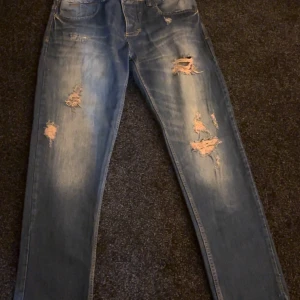 Slim fit blå jeans från Quest, stl 33 - Snygga slim fit jeans från Quest i klassisk blå tvätt med slitna och rivna detaljer på både fram- och baksidan. Modellen har fem fickor, normal midja och raka ben. Perfekt för dig som gillar en avslappnad streetstil.