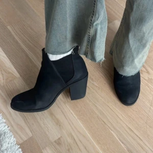 Svarta stövletter med klack - Säljer ett par svarta stövletter med rund tå och stabil hög klack. Skorna har en stilren design och är tillverkade i mocka, vilket ger dem en mjuk och matt finish. Perfekta att matcha med jeans eller kjol för en cool look.