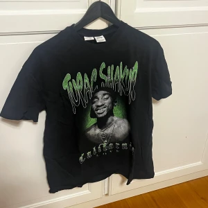 Svart 2PAC t-shirt från Bershka - Cool svart t-shirt från Bershka med stort 2PAC-tryck och texten 'California Love' i grönt och vitt på framsidan. Klassisk rund hals och korta ärmar. Materialet är mjuk bomull, perfekt för dig som gillar streetwear och ikoniska prints.