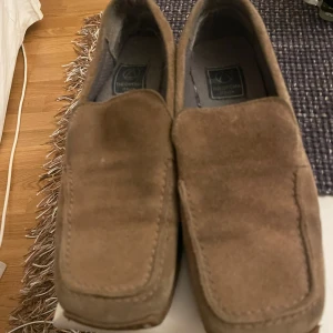 Bruna mockaloafers från Theorema - Snygga bruna loafers från Theorema, tillverkade i mjuk mocka. Skorna har en fyrkantig tå och klassisk slip-on design utan snörning. Perfekta för dig som gillar en stilren och avslappnad look med italiensk känsla.