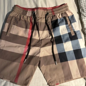 Rutig Burberry shorts med snörning - Snygga Burberry shorts med klassiskt rutigt mönster i beige, svart, blått och rött. De har elastisk midja med svart snörning och fickor både fram och bak. Perfekt för dig som vill ha en clean och lyxig vibe i sommar. Materialet känns lätt och skönt mot huden.