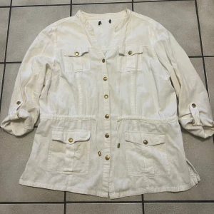 Vit overshirt med guldknappar storlek L/XL - Stilren vit overshirt med guldiga knappar och fyra fickor framtill. Jackan har dragsko i midjan för snygg passform och ärmarna kan rullas upp och fästas med knapp. Perfekt för lager-på-lager och en clean look.