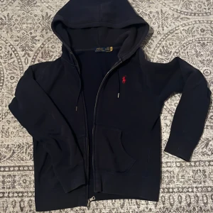Svart hoodie från Polo Ralph Lauren XS - Svart hoodie från Polo Ralph Lauren i storlek XS. Tröjan har huva med snörning, dragkedja framtill och fickor. Klassisk röd broderad logga på bröstet. Perfekt för chill dagar och streetwear. Mjuk bomullskvalitet och långärmad modell.