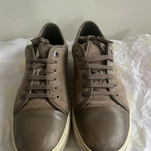 Bruna sneakers från Lanvin i mocka - Snygga bruna sneakers från Lanvin med ovandel i mocka och tåparti i skinn. Skorna har rund tå, snörning och vit platt sula. Perfekta för dig som gillar stilrena och exklusiva sneakers med en lyxig känsla. Dom ända slitagen som finns är lite på captoen där färgen har gått bort samt på framsidan av skon. Sulan har blivit lite missfärgad men det går enkelt att fixa hos en skomakare