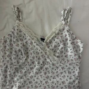 Blommig vit linnetopp med spets från SHEIN - Supersöt linnetopp från SHEIN i vit med små rosa blommor och gröna blad. Toppen har ribbad struktur, spetsdetaljer längs v-ringad hals och justerbara axelband med knyt. Aldrig använd