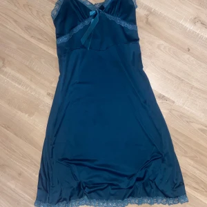 Marinblå långklänning med spets - Snygg marinblå långklänning med tunna axelband och v-ringning. Klänningen har spetsdetaljer längs halsringningen och under bysten samt en söt rosett framtill. Materialet är mjukt och följsamt. 