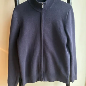 Mörkblå kofta med dragkedja från Bläck - Säljer en stilren mörkblå kofta från Bläck i storlek S. Koftan har hel dragkedja framtill, ribbad krage och är tillverkad i ett mjukt stickat material. Perfekt för lager-på-lager och enkel att matcha med det mesta. Nypris 800