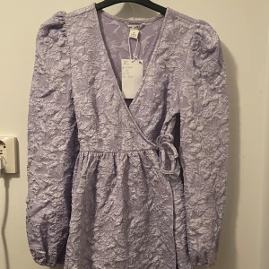 Ljuslila omlottklänning från Monki - Superfin ljuslila omlottklänning från Monki i storlek XS. Klänningen har puffiga långärmar, v-ringning och ett vackert blommigt mönster i strukturerat tyg. Knytband i midjan ger en snygg siluett och den är perfekt för dig som gillar detaljer.