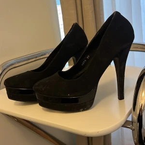 Svarta pumps med hög klack och platå - Snygga svarta pumps från Elle Woman med rund tå, hög klack och platå. Skorna har en stilren design i mocka-look och passar perfekt när du vill höja din outfit. Klacken är smal och ger en elegant siluett. Storlek 37.