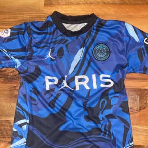 PSG Mbappé blå kortärmad matchtröja - Snygg PSG fotbollströja med Mbappé och nummer 7 på ryggen. Tröjan är blå med svarta och ljusblå abstrakta mönster, korta ärmar och svart krage. Officiella Ligue 1 och Jordan-loggor på bröstet, tillverkad i lätt polyester som andas.