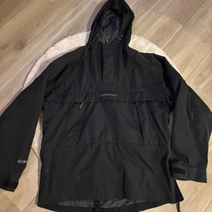 Svart anorak jacka från Everest M - Svart vindjacka från Everest med GoldFlexSystem, storlek M. Jackan har huva med snörning, stora fickor framtill och justerbara ärmslut med kardborre. Perfekt för blåsiga dagar och outdoor-aktiviteter. Snygg och funktionell design med diskret logga på bröstet.