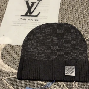 Louis Vuitton svart rutig mössa - Svart mössa från Louis Vuitton med diskret rutigt mönster och ribbad kant. Märkesplakett i metall framtill ger en lyxig touch. Perfekt accessoar för dig som vill ha en stilren och exklusiv look under kyliga dagar.
