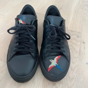 Svarta Axel Arigato sneakers med broderad fågel - Säljer mina svarta Axel Arigato-skor eftersom de tyvärr är för små för mig. Storlek 41. Skorna är i mycket bra skick och har inga defekter. Snygg design med broderade fåglar på sidorna och framtill.  Frågor eller bud är varmt välkomna – hör gärna av dig om du är intresserad! 